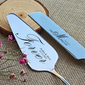 Könnte beinhalten: Silbernes Tortenheber- und Messerset. Der Tortenheber trägt das Wort "Forever" in Schreibschrift sowie Namen und ein Datum. Das Messer ist mit "The Morgans" und einem Datum graviert. Rosa Blumen und eine Holzscheibe im Hintergrund.