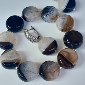 Puede incluir: Un conjunto de collar y pendientes con grandes cuentas redondas de ágata en tonos negro, marrón y blanco. Las cuentas están ensartadas con pequeños conectores plateados. También se ve un pendiente plateado con un diseño texturizado.