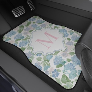 May include: Car floor mat with a floral pattern of blue and green hydrangeas. The mat features a white central design with a pink letter "M" inside a decorative frame. The mat is placed in a car.