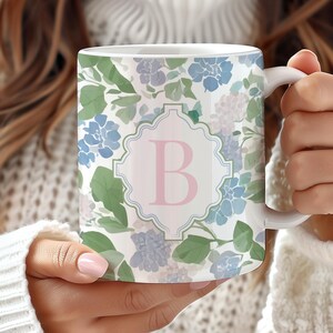 Op de afbeelding: Witte keramische mok met een bloemenpatroon van blauwe, groene en lavendelkleurige hortensia's. Een roze letter "B" is gecentreerd in een decoratief frame. De mok heeft een wit oor en wordt vastgehouden door een persoon die een witte trui draagt.