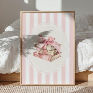 May include: A framed print featuring a stack of books tied with a pink ribbon, set against a pink and white striped background. The books are illustrated in watercolor style, with a vintage aesthetic. The print is framed in a light wood frame.