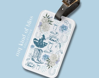 May include: White luggage tag with a blue and white design featuring cowboy boots, horseshoes, flowers, and the text "yee haw". The tag is attached to a black leather strap.
