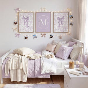 Puede incluir: Una escena de dormitorio con una cama blanca adornada con almohadas en tonos rosa y lavanda. Encima de la cama, tres cuadros enmarcados con diseños de lazo y la letra "M". Un cárdigan color crema y un libro descansan sobre la cama.