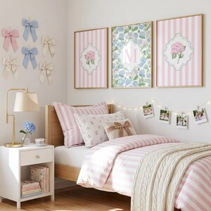 May include: A bedroom interior featuring a bed with pink and white striped bedding and floral cushions. Above the bed are three framed artworks. A white bedside table with a gold lamp and books is on the left. Decorative bows are attached to the wall.
