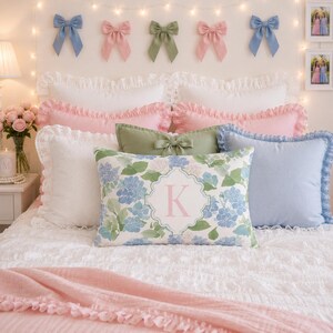 May include: A bed with multiple pillows and decorative bows. The pillows are white, pink, and blue, with floral and solid patterns. The bows are blue, pink, and green. The bed is covered with a white, textured bedspread and a pink blanket.