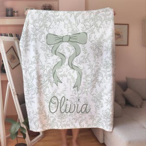 My Personalized Sage Green Bow Toile Name Blanket features a classic olive toile print with delicate flowers and birds on a crisp white background, topped with a big coquette-style bow and your custom name in an elegant script below. Soft feminine and beautifully detailed, it’s perfect for babies, kids, teens and adults. Great for sleepover favors, hospital or coming-home blankets, announcement photos, dorm rooms, teen bedrooms, Christmas, birthdays, sisters, newborn keepsakes and new parents