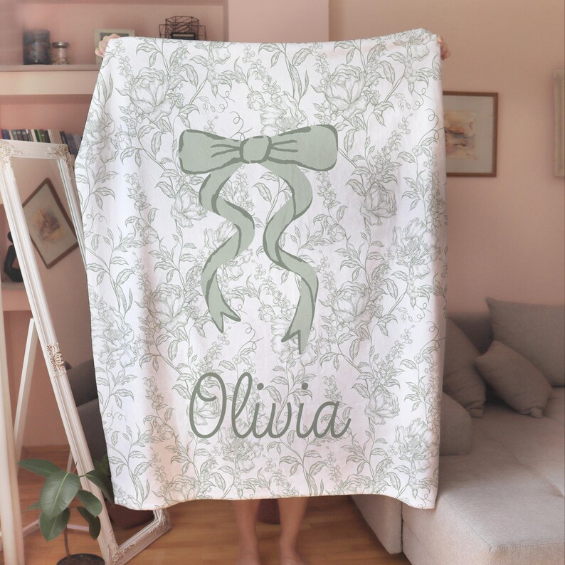 My Personalized Sage Green Bow Toile Name Blanket features a classic olive toile print with delicate flowers and birds on a crisp white background, topped with a big coquette-style bow and your custom name in an elegant script below. Soft feminine and beautifully detailed, it’s perfect for babies, kids, teens and adults. Great for sleepover favors, hospital or coming-home blankets, announcement photos, dorm rooms, teen bedrooms, Christmas, birthdays, sisters, newborn keepsakes and new parents