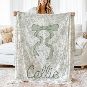 My Personalized Sage Green Bow Toile Name Blanket features a classic olive toile print with delicate flowers and birds on a crisp white background, topped with a big coquette-style bow and your custom name in an elegant script below. Soft feminine and beautifully detailed, it’s perfect for babies, kids, teens and adults. Great for sleepover favors, hospital or coming-home blankets, announcement photos, dorm rooms, teen bedrooms, Christmas, birthdays, sisters, newborn keepsakes and new parents