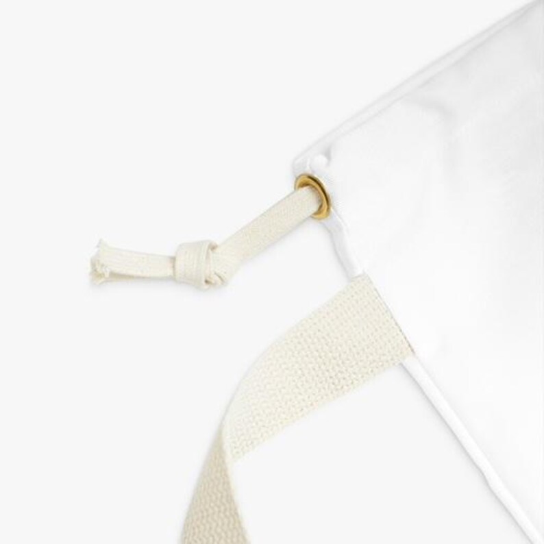 May include: A close-up of a white fabric bag with a cream drawstring and strap. The drawstring is knotted, and a gold-coloured grommet reinforces the bag's opening. The strap is made of a woven, cream-coloured material.