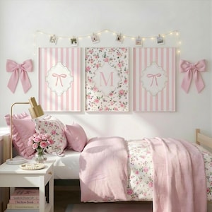 Puede incluir: Una escena de dormitorio con una cama blanca, ropa de cama rosa y almohadas con estampado floral. Tres obras de arte enmarcadas con rayas rosas y diseños florales cuelgan sobre la cama. Lazos rosas adornan la pared y una lámpara dorada se encuentra en una mesita de noche blanca.