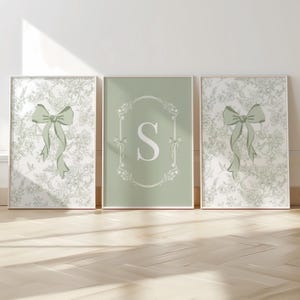 May include: Three framed art prints. Two prints feature a green bow on a white floral background. The center print is sage green with a white letter "S" inside a decorative frame. The prints are displayed on a light wood floor.