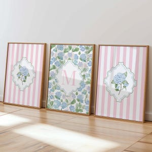 May include: Three framed wall art prints in light wood frames. The two outer prints feature vertical pink and white stripes with a central white quatrefoil emblem containing a blue hydrangea illustration. The center print shows a blue hydrangea pattern with a central white quatrefoil emblem featuring a large pink letter M.