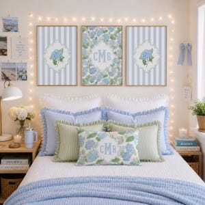 Könnte beinhalten: Drei gerahmte Wandbilder mit blauen Hortensien-Illustrationen und einem zentralen Monogramm-Design auf gestreiftem Hintergrund, platziert über einem Bett mit blau-weißer Bettwäsche, inklusive eines Zierkissens mit dem Monogramm CMB.