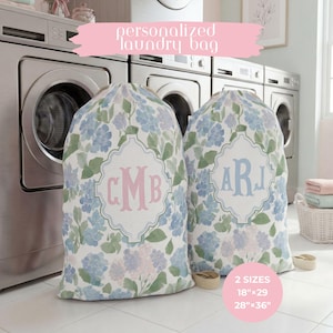 May include: Two fabric laundry bags featuring a watercolor floral pattern in shades of blue, green, and white. Each bag displays a central decorative frame with personalized monogram letters in pink or blue. Text includes 2 SIZES 18"x29 28"x36".