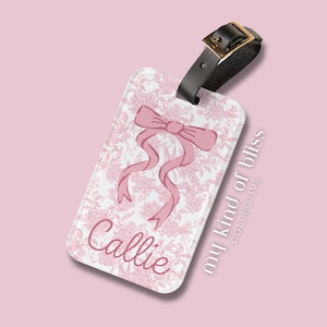 May include: A rectangular luggage tag with a pink floral pattern and a pink bow design. The name "Callie" is written in cursive. The tag has a black leather strap with a gold buckle.