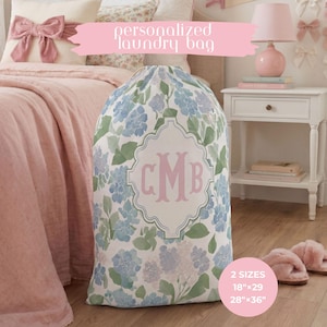 May include: A laundry bag featuring a white background with a blue and green hydrangea floral pattern. A central decorative frame contains the pink monogram letters CMB. The bag is available in two sizes: 18 inches by 29 inches and 28 inches by 36 inches.