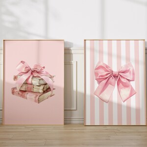 My cute coquette set of three pink bow prints. The bows have a watercolour look to the. Two prints have white and pink stripes in the background with a large bow in front. The middle print has a book stack on a pink background with a bow. Nursery Art