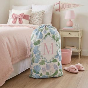 Personalized hydrangea monogram laundry bag styled in a pink coquette-inspired teen bedroom beside a bed with blush bedding, bow pillow, pink lamp, slippers, and nightstand, cute dorm room storage gift for girls.