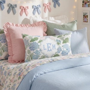 May include: A rectangular throw pillow featuring a blue and green floral pattern with a central white scalloped frame containing the monogram LEM in light blue letters, positioned among various solid and patterned pillows on a bed.