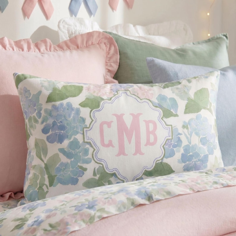Close-up of custom hydrangea monogram lumbar pillow with pastel blue hydrangeas, soft pink initials, and feminine bedroom styling.