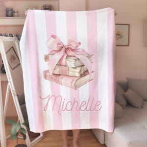 My Custom Name Book Lover Coquette Blanket. Featuring a pink and white striped background with a watercolor book stack wrapped in a bow, this plush blanket is as cozy as it is cute. Add your custom name underneath for a personalized, preppy, coquette touch. Perfect for book lovers, girls bedrooms, baby showers, newborns, or a stylish preppy touch to dorm