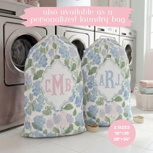 Op de afbeelding: Twee stoffen waszakken met een aquarel bloemenpatroon in tinten blauw, groen en lichtpaars. Elke zak heeft een centraal wit decoratief kader met een gepersonaliseerd monogram in roze of blauwe letters. Tekst: 46cmx74cm en 71cmx91cm.