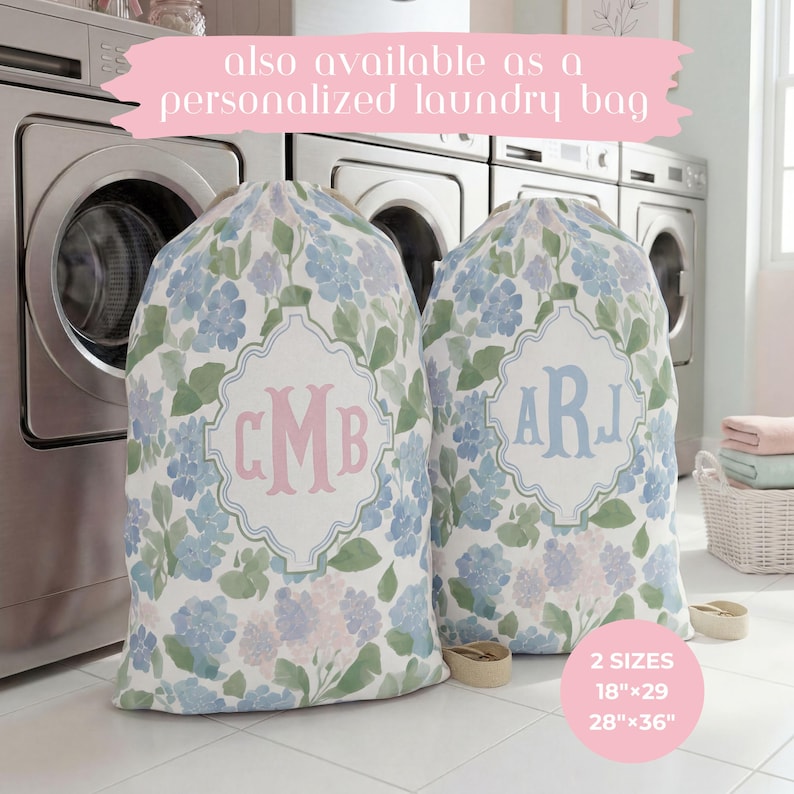 Op de afbeelding: Twee stoffen waszakken met een aquarel bloemenpatroon in tinten blauw, groen en lichtpaars. Elke zak heeft een centraal wit decoratief kader met een gepersonaliseerd monogram in roze of blauwe letters. Tekst: 46cmx74cm en 71cmx91cm.