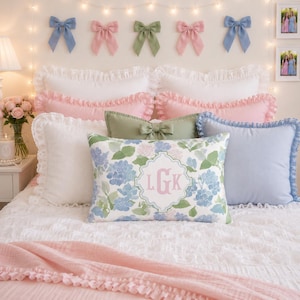 May include: A collection of decorative pillows on a bed, including a rectangular pillow with a blue and green floral pattern and the monogram LGK in the center, alongside solid white, light blue, and sage green pillows with ruffled edges.