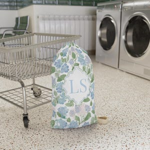 Custom monogram floral laundry bag standing beside rolling laundry cart in dorm laundry room, preppy dorm essential