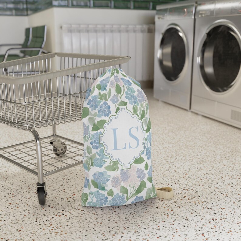 Custom monogram floral laundry bag standing beside rolling laundry cart in dorm laundry room, preppy dorm essential