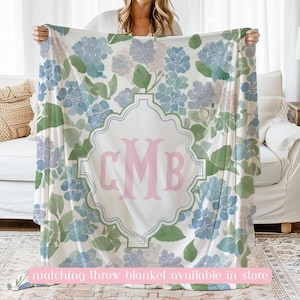 Op de afbeelding: Een witte plaid met een herhalend patroon van blauwe en paarse hortensiabloemen met groene bladeren. In het midden omsluit een decoratief lichtblauw kader de grote roze monogramletters CMB in een schreeflettertype.