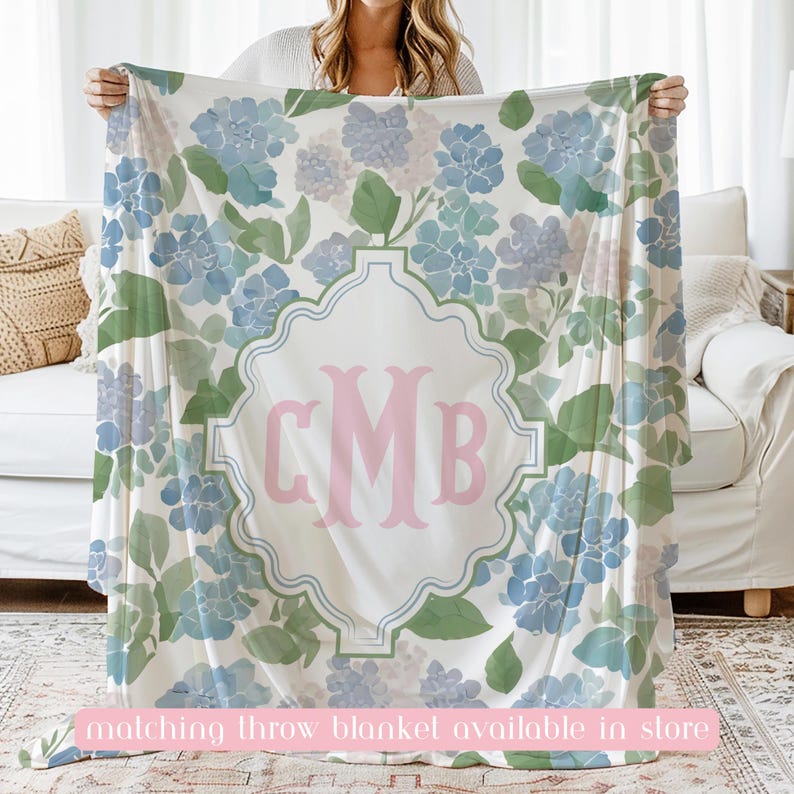Op de afbeelding: Een witte plaid met een herhalend patroon van blauwe en paarse hortensiabloemen met groene bladeren. In het midden omsluit een decoratief lichtblauw kader de grote roze monogramletters CMB in een schreeflettertype.