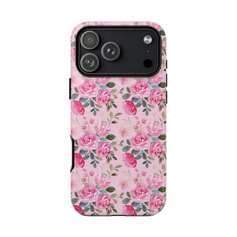 Pu&ograve; includere: Una custodia per smartphone con un motivo ripetuto di rose rosa e foglie verdi su sfondo rosa chiaro. La custodia presenta un ritaglio nero per il modulo fotocamera nella parte superiore, che rivela tre lenti circolari e un flash del dispositivo.