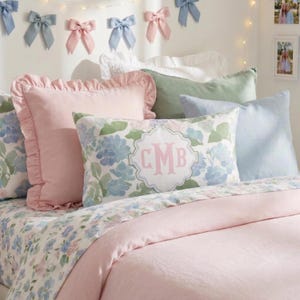 May include: A bed with a light pink comforter and floral patterned sheets. Several pillows in shades of pink, blue, and green are arranged on the bed. A pillow with the initials "CMB" is visible. Decorative bows in pink and blue adorn the wall.