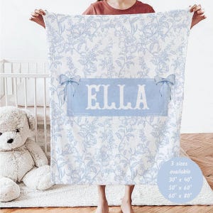 May include: A white and blue floral patterned blanket with the name "ELLA" in a light blue banner. The blanket has a bow on each side of the banner. Available in 3 sizes: 30" x 40", 50" x 60", and 60" x 80".