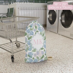 Small personalized hydrangea laundry bag with blue initials beside laundry cart in laundromat, cute dorm room essential