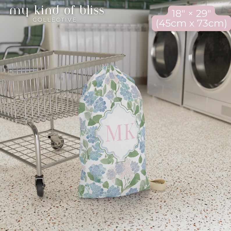 Small personalized hydrangea laundry bag with blue initials beside laundry cart in laundromat, cute dorm room essential