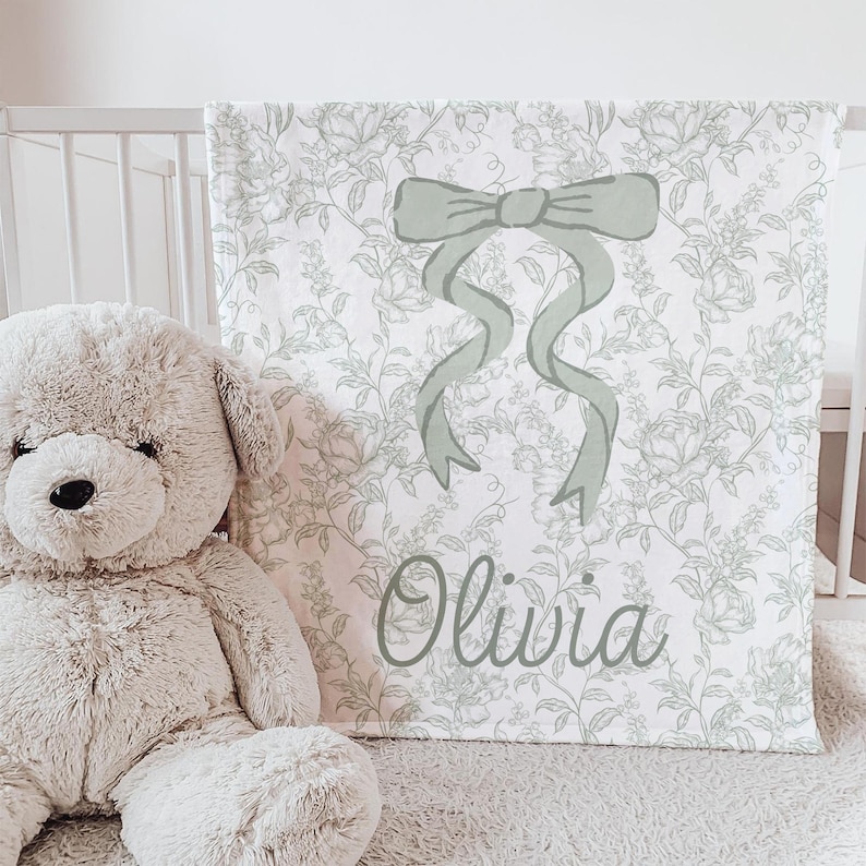 My Personalized Sage Green Bow Toile Name Blanket features a classic olive toile print with delicate flowers and birds on a crisp white background, topped with a big coquette-style bow and your custom name in an elegant script below. Soft feminine and beautifully detailed, it’s perfect for babies, kids, teens and adults. Great for sleepover favors, hospital or coming-home blankets, announcement photos, dorm rooms, teen bedrooms, Christmas, birthdays, sisters, newborn keepsakes and new parents