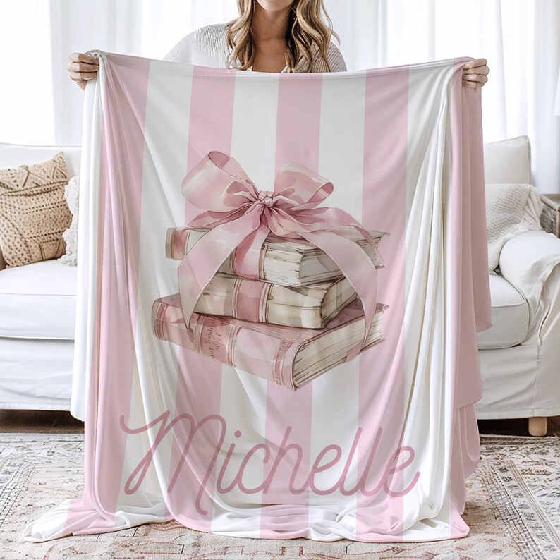 My Custom Name Book Lover Coquette Blanket. Featuring a pink and white striped background with a watercolor book stack wrapped in a bow, this plush blanket is as cozy as it is cute. Add your custom name underneath for a personalized, preppy, coquette touch. Perfect for book lovers, girls bedrooms, baby showers, newborns, or a stylish preppy touch to dorm bedrooms. Whether its for a teen, college student, daughter, roommate, friend or newborn baby, its a cute and cozy addition to any space.