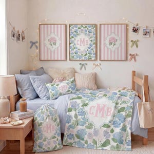 Op de afbeelding: Een slaapkamerset met een blauw en roze bloemendeken, een bijpassende waszak en een sierkussen, allemaal voorzien van het monogram CMB. Drie ingelijste wandplaten met roze strepen en bloemmotieven hangen boven het bed.