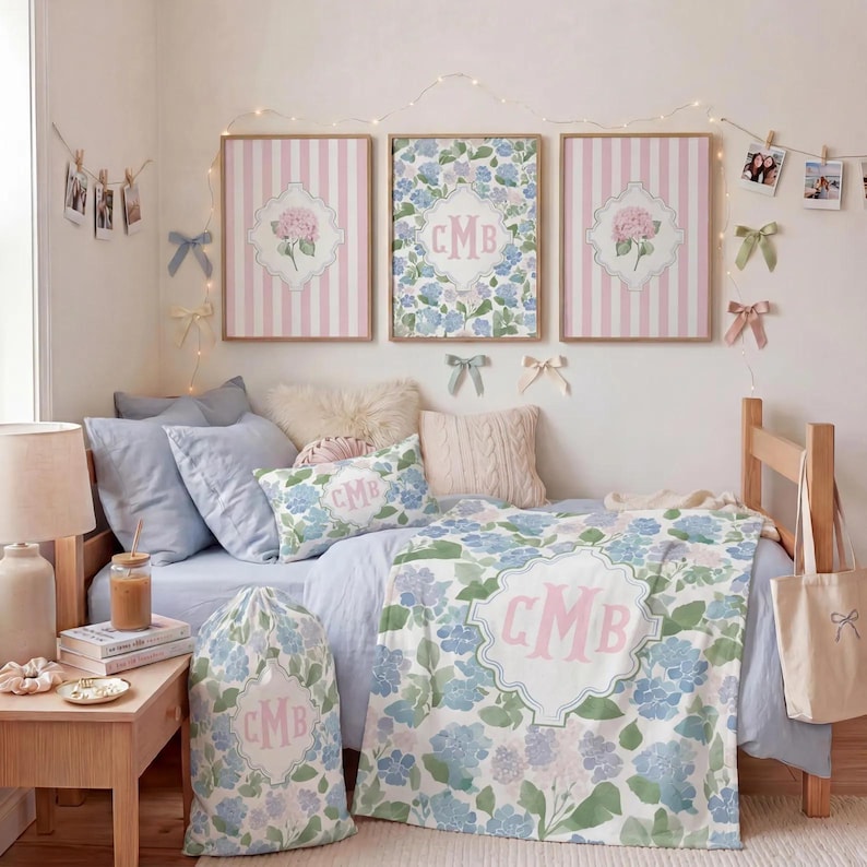 Op de afbeelding: Een slaapkamerset met een blauw en roze bloemendeken, een bijpassende waszak en een sierkussen, allemaal voorzien van het monogram CMB. Drie ingelijste wandplaten met roze strepen en bloemmotieven hangen boven het bed.