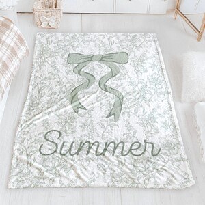 My Personalized Sage Green Bow Toile Name Blanket features a classic olive toile print with delicate flowers and birds on a crisp white background, topped with a big coquette-style bow and your custom name in an elegant script below. Soft feminine and beautifully detailed, it’s perfect for babies, kids, teens and adults. Great for sleepover favors, hospital or coming-home blankets, announcement photos, dorm rooms, teen bedrooms, Christmas, birthdays, sisters, newborn keepsakes and new parents