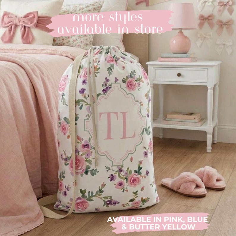 May include: A white laundry bag with a floral pattern of pink roses and purple flowers. The bag has a pink monogram and a beige shoulder strap. Pink fuzzy slippers are on the floor. The text "more styles available in store" is visible.