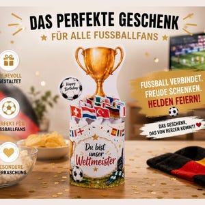 Könnte beinhalten: Eine dekorative Geschenkbox in Form einer Trophäe mit einer goldenen Tasse oben, umgeben von Flaggen und Fußbällen. Die Box enthält deutschen Text, darunter "Das Perfekte Geschenk" und "Du bist unser Weltmeister".