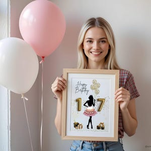 May include: A framed print with the words "Happy Birthday 17" and a silhouette of a person in a pink skirt. The print is held by a person, with two balloons in the background. One balloon is pink, and the other is white.