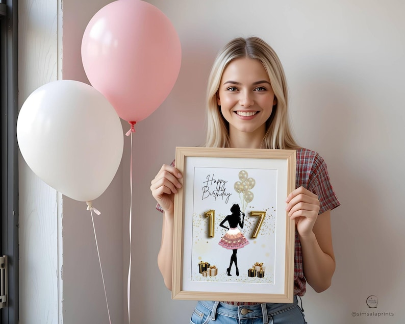 May include: A framed print with the words "Happy Birthday 17" and a silhouette of a person in a pink skirt. The print is held by a person, with two balloons in the background. One balloon is pink, and the other is white.