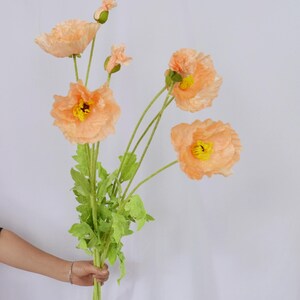 May include: A bouquet of artificial peach-colored poppy flowers with yellow centers and green stems and leaves. The flowers are in various stages of bloom, with some buds visible. The bouquet is held by a person.
