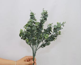 May include: A bundle of artificial eucalyptus branches with round, green leaves. The leaves vary in shades of green, and the stems are a darker green. The branches are held by a hand against a white background.