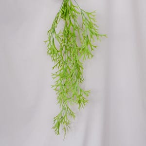 Op de afbeelding: Een heldergroene, kunstmatige plantenstengel met fijne, varenachtige bladeren. De plant hangt tegen een effen witte achtergrond, wat de levendige kleur en gedetailleerde textuur benadrukt. Geschikt voor interieurdecoratie.