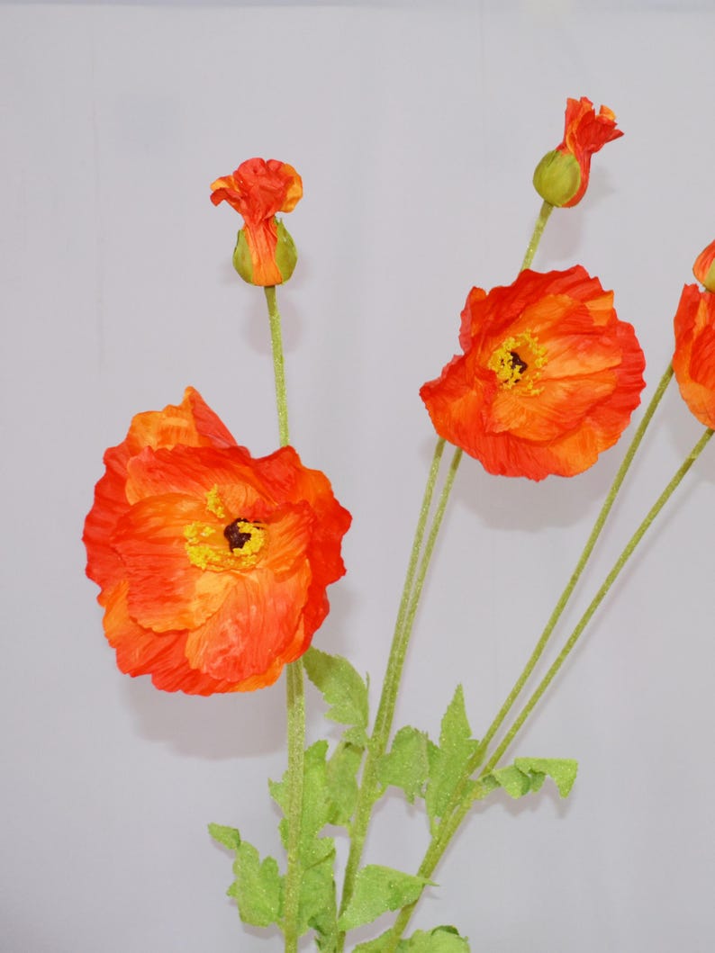 May include: Close-up of orange and yellow artificial poppy flowers with green stems and leaves. The flowers are in various stages of bloom, with some fully open and others still in bud form. The petals have a textured appearance.